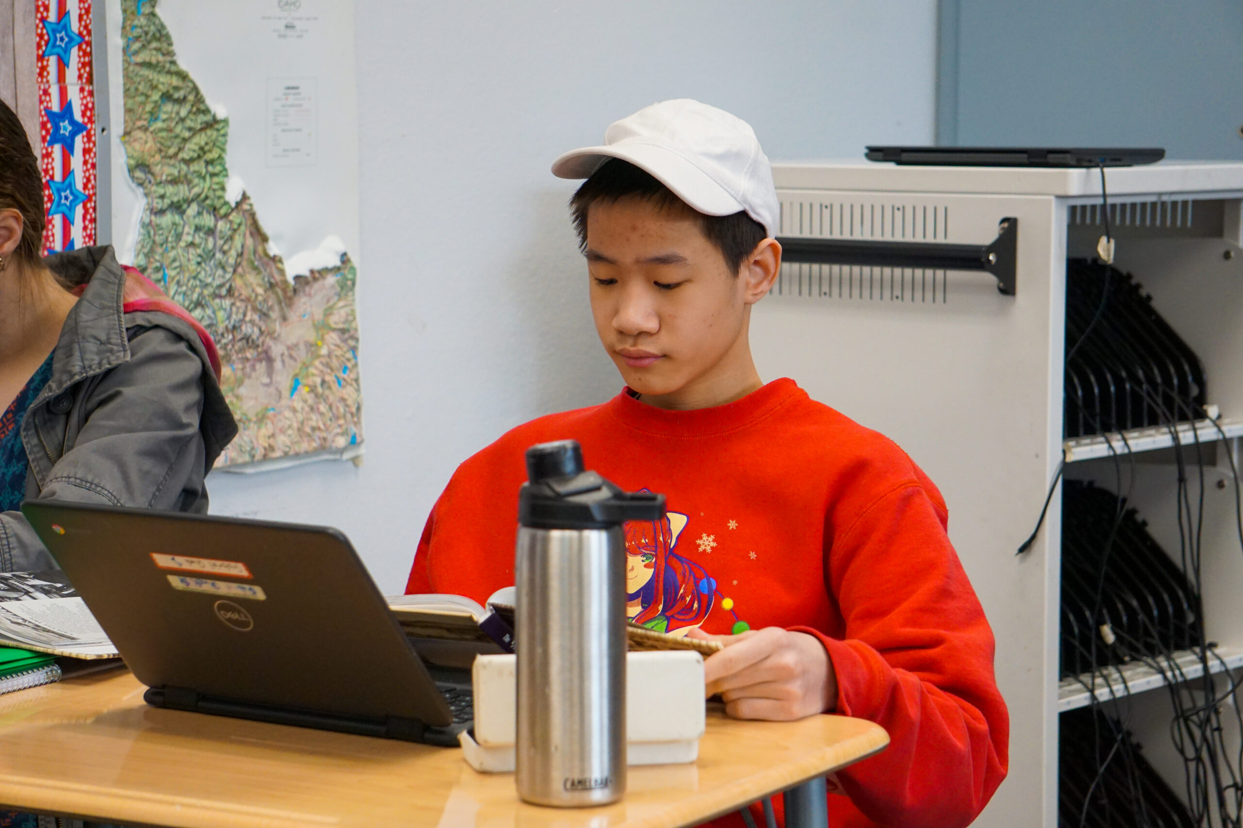 Student in red sweatshirt and white baseball cap using laptop while at a desk
